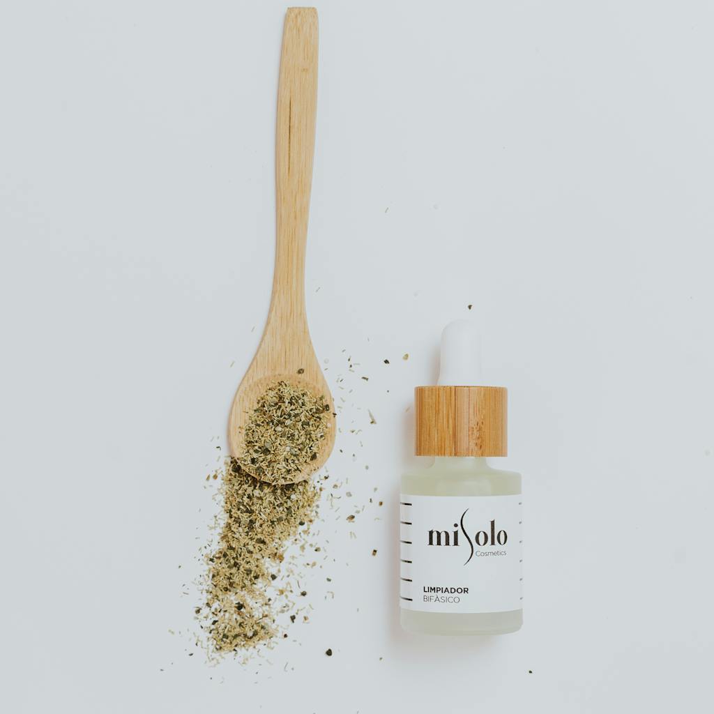 Flat lay of a natural skincare bottle with herbs and wooden spoon on a white background.