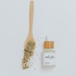 Flat lay of a natural skincare bottle with herbs and wooden spoon on a white background.
